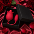 A red rose-shaped intimate massager with a matching applicator inside an open black gift box, set on red satin fabric with red roses and rose petals around it.